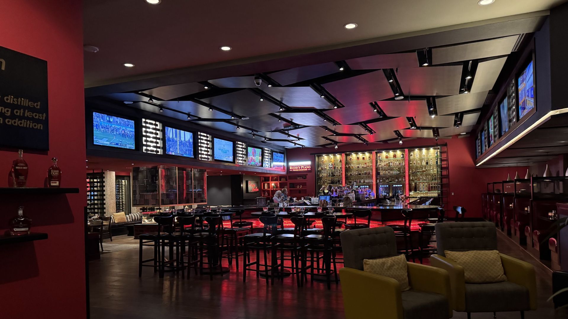 Sports bar interior with multiple TVs, bar seating, red and black decor, and dim lighting.