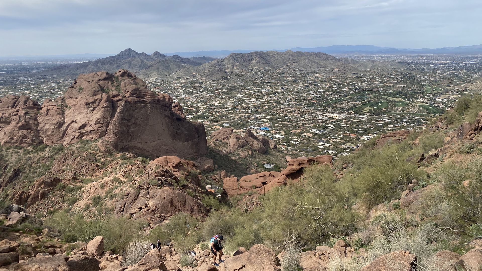 Hiker on a rocky trail overlooking a sprawling city, mountains in the distance. Cloudy sky.