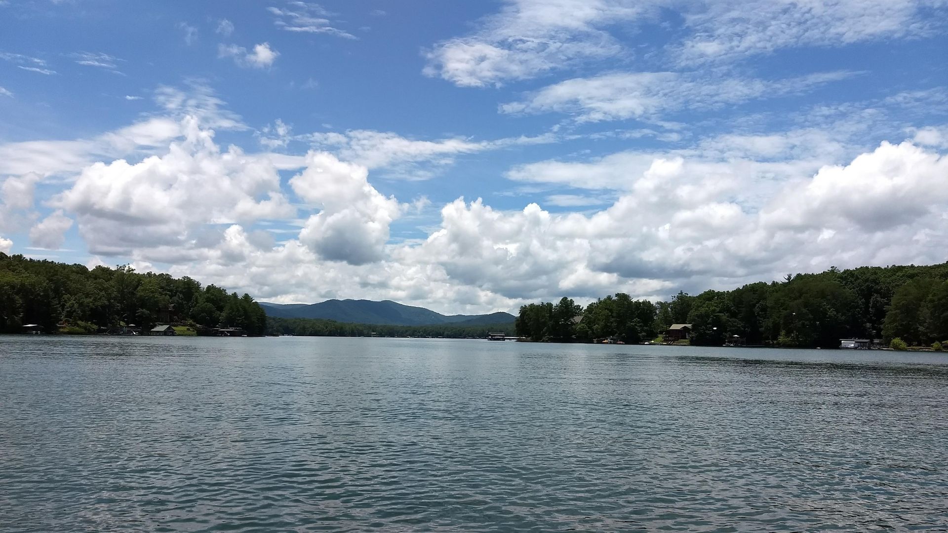 A wide, calm lake surrounded by dense green forests under a bright blue sky filled with large, fluffy white clouds.