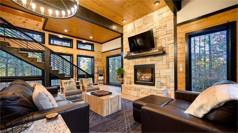 Cozy cabin living room with fireplace, leather furniture, and large windows overlooking trees.