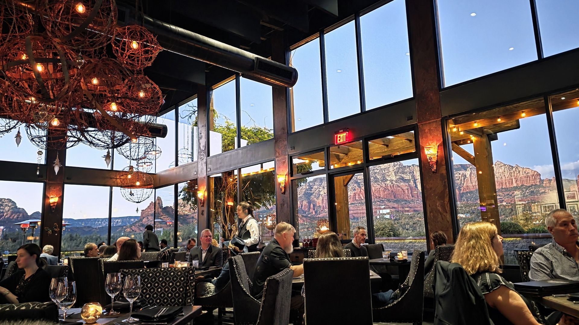 Restaurant interior with large windows overlooking a red rock landscape. Diners seated at tables, chandeliers.
