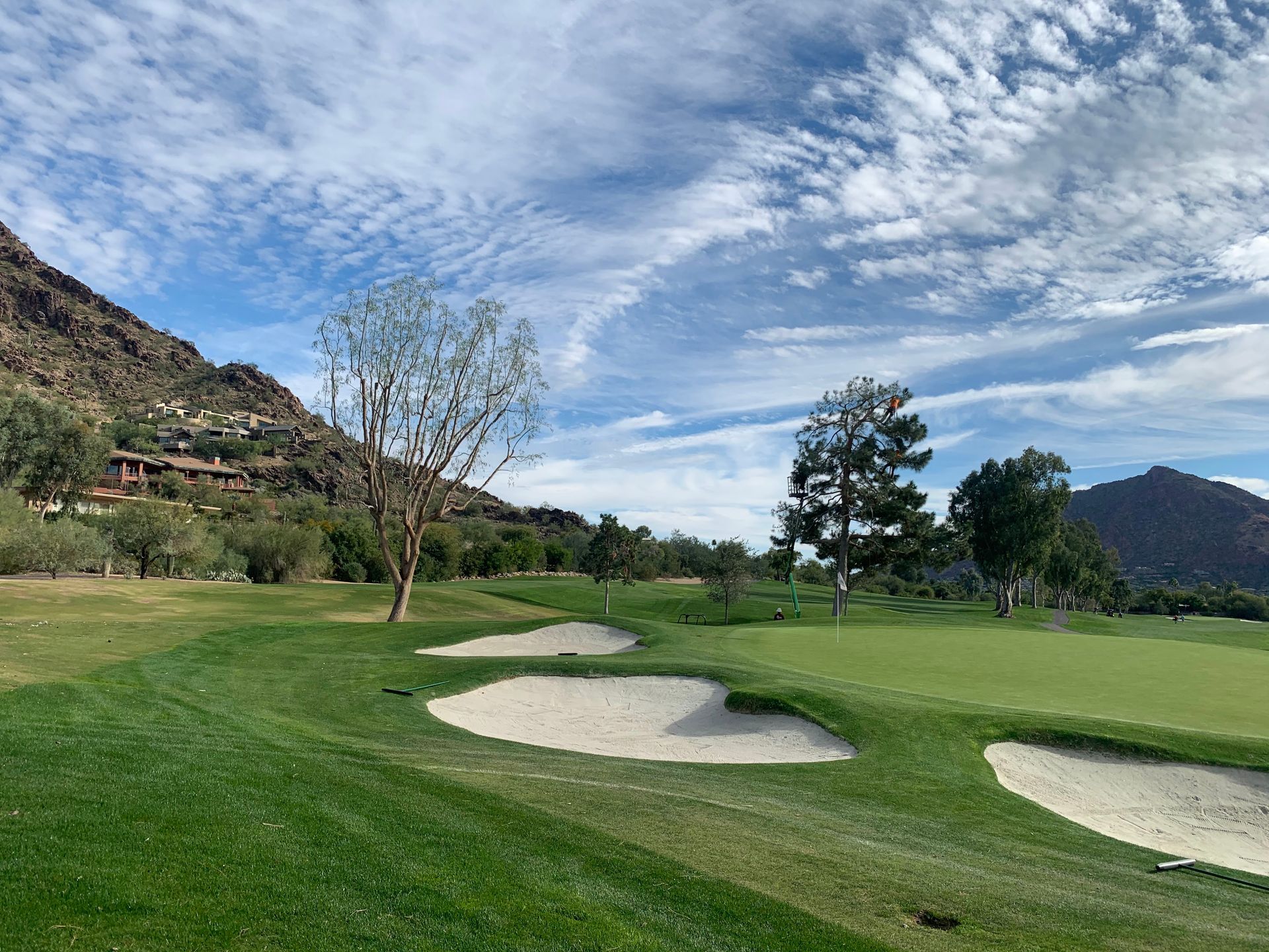 Golf course scene with green grass, sand traps, trees, and mountains under a cloudy sky.