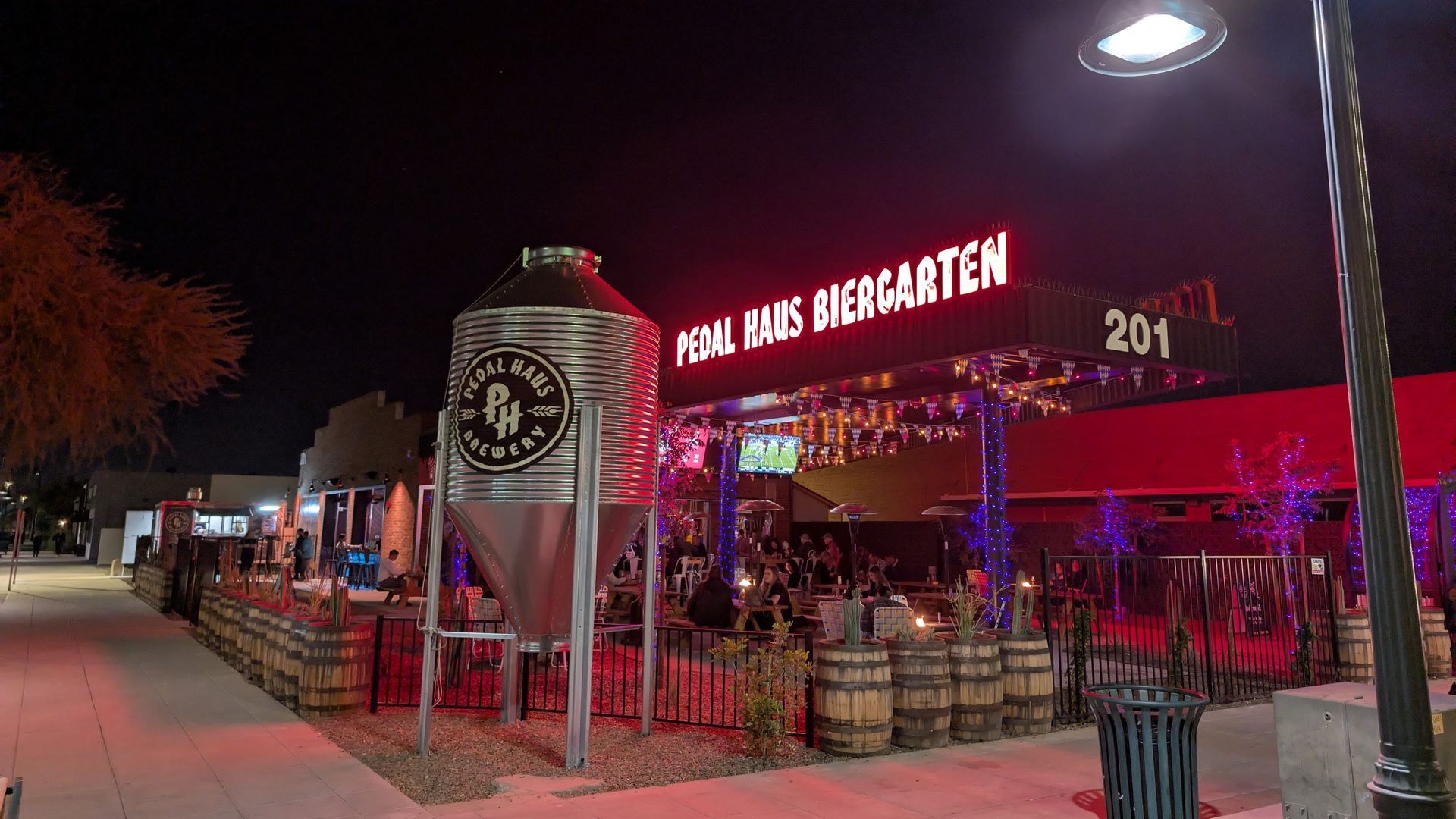 Night scene of a biergarten: red neon sign, large metal tank, outdoor seating, barrels, and people.