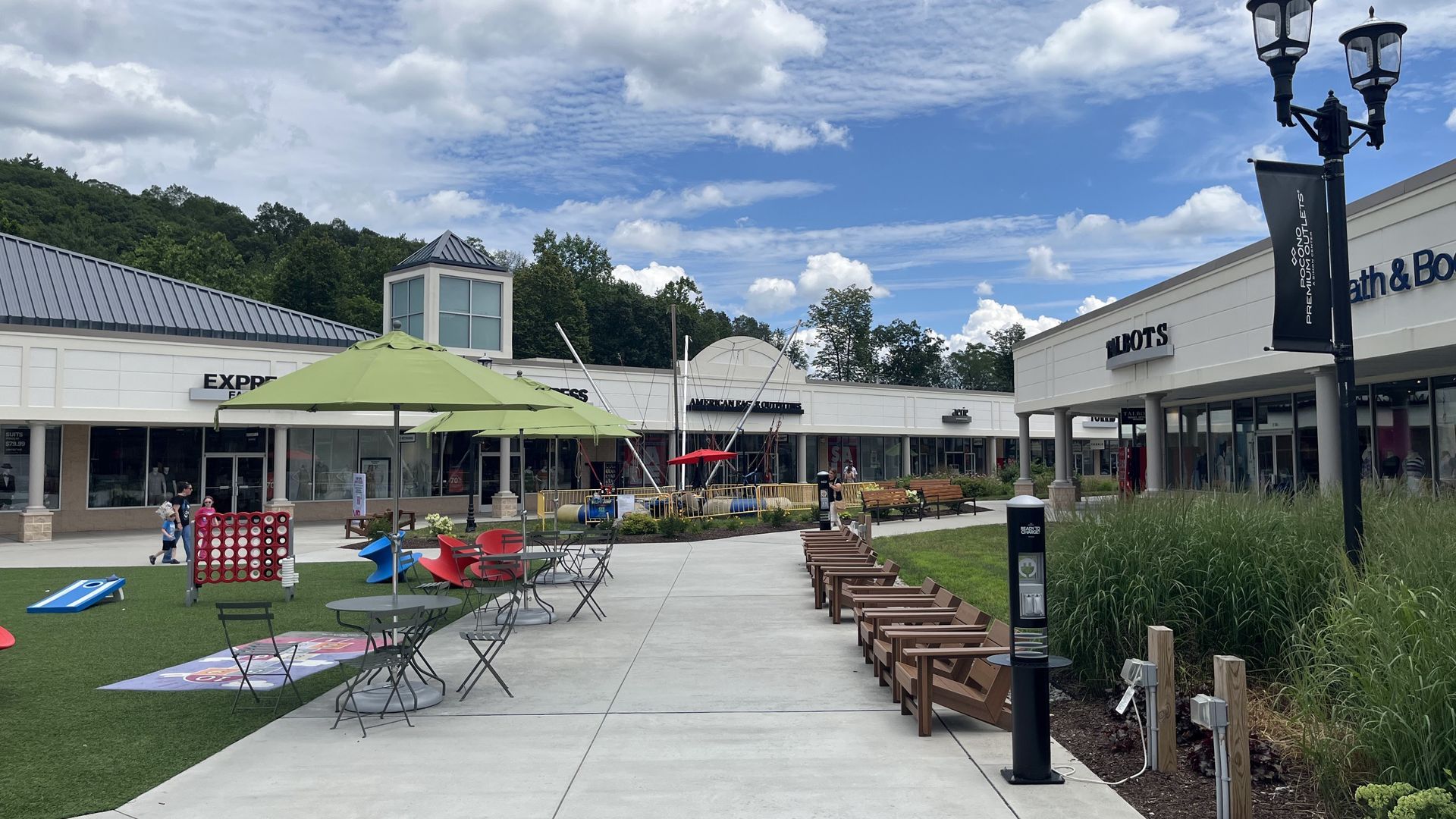 An outdoor shopping center plaza with a grassy lawn, cafe tables under umbrellas, and storefronts under a blue sky.