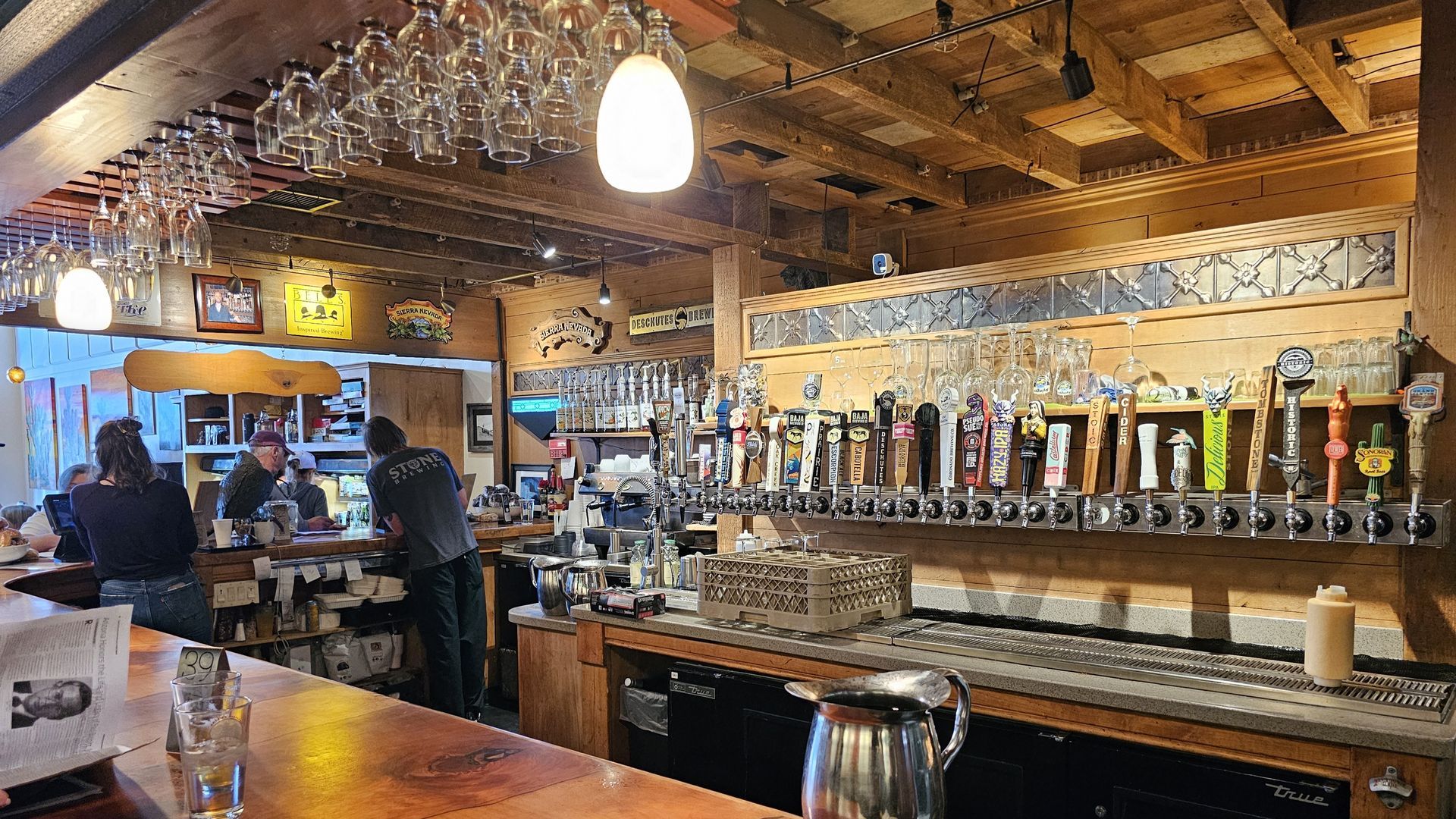Bar interior with beer taps, hanging glasses, and customers at the bar. Wooden decor, warm lighting.