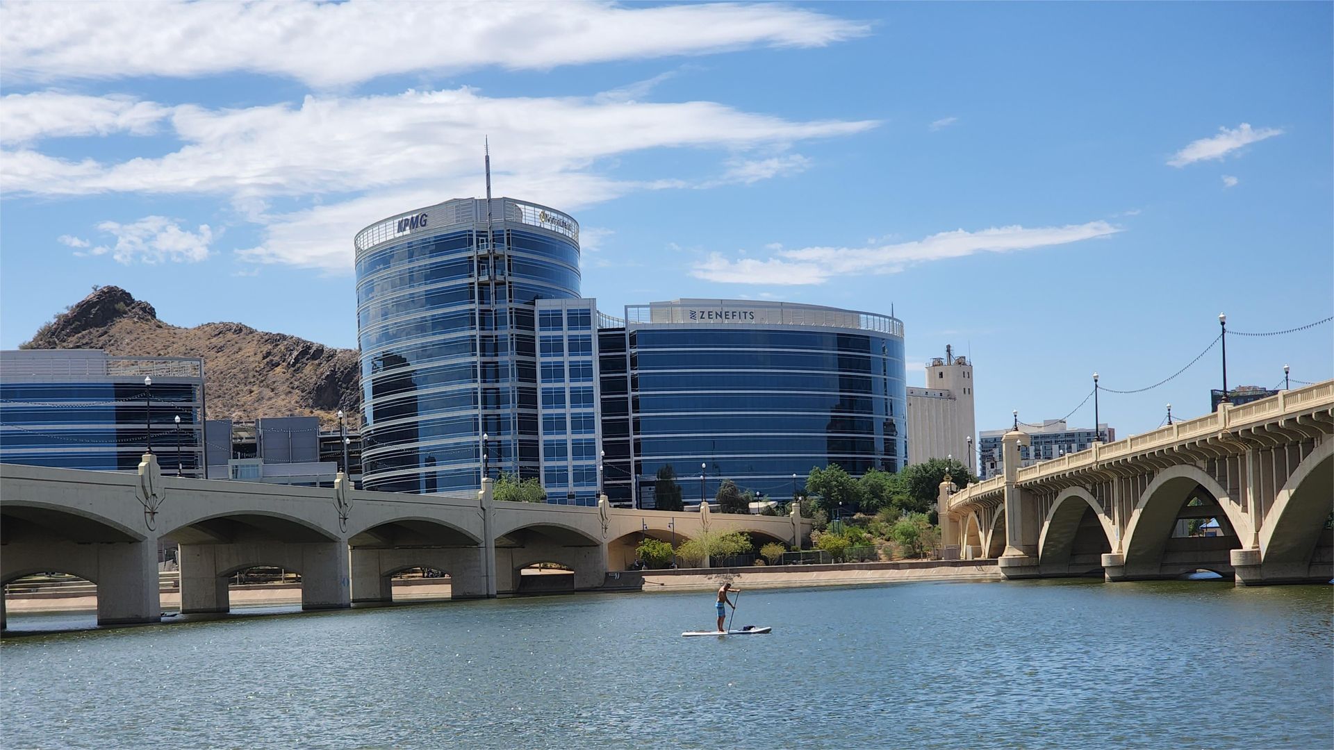 Modern buildings, stone bridge, and mountains reflected in a lake on a sunny day.