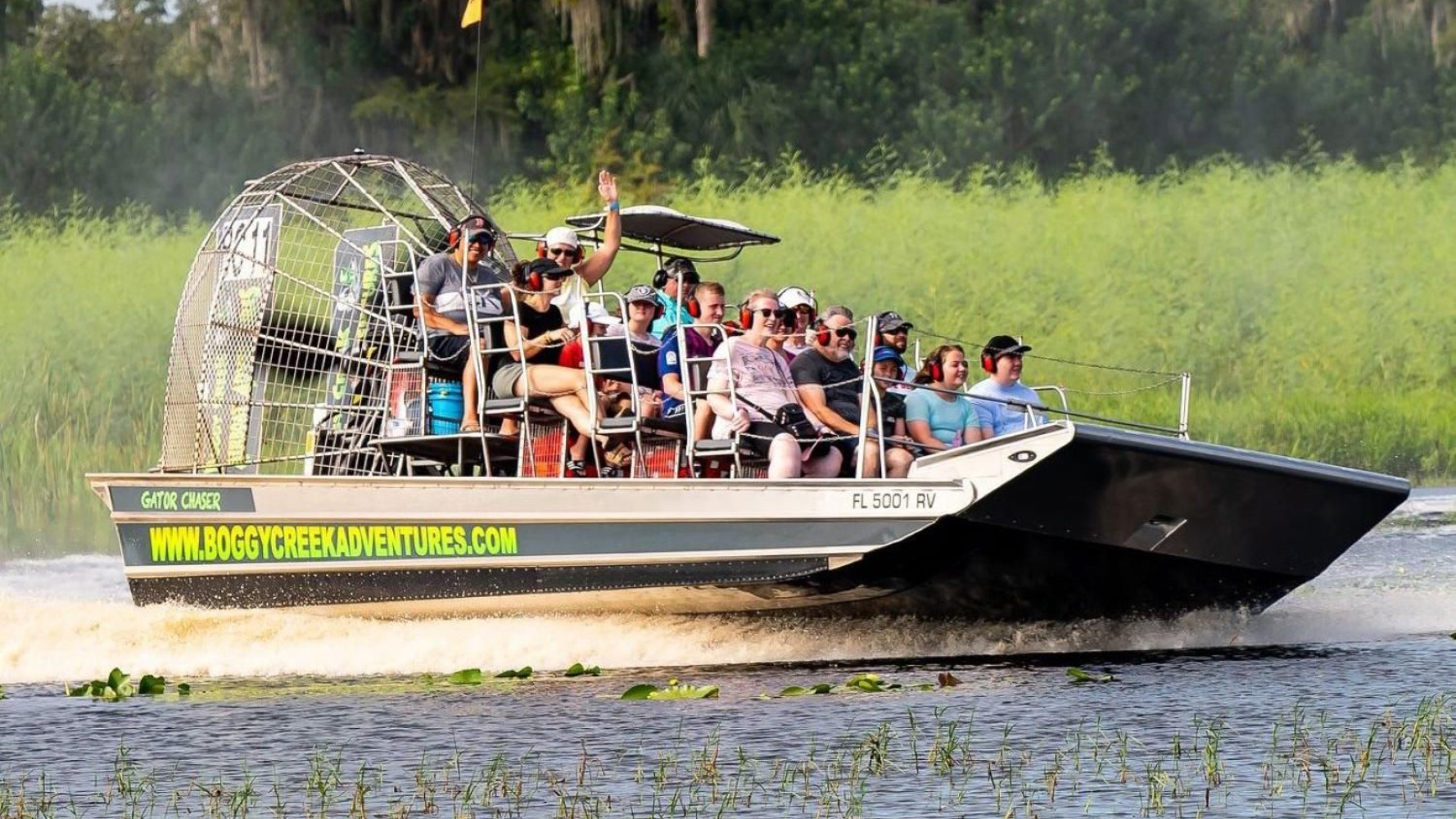 Airboat with passengers on a swamp tour, splashing water, waving, sunny day.