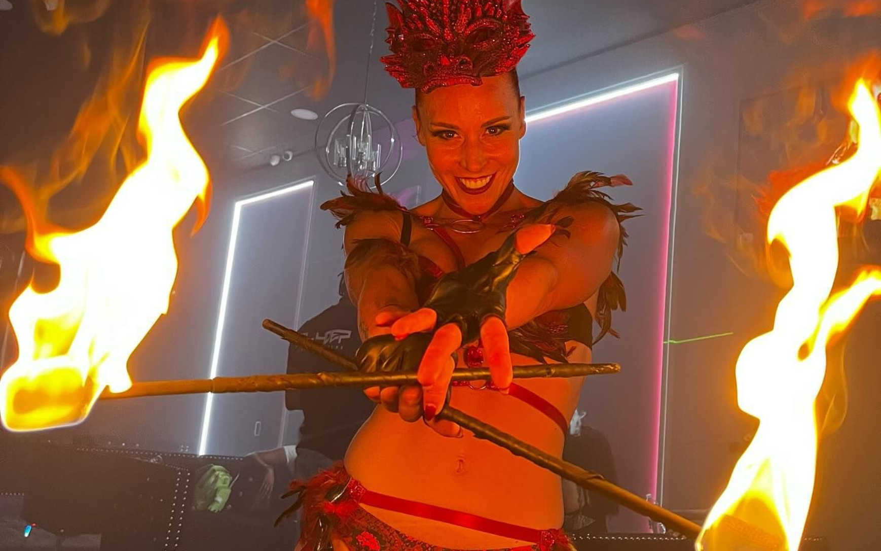 Woman holding flaming rods, smiling, with a red-accented costume. Indoor, stage lighting.