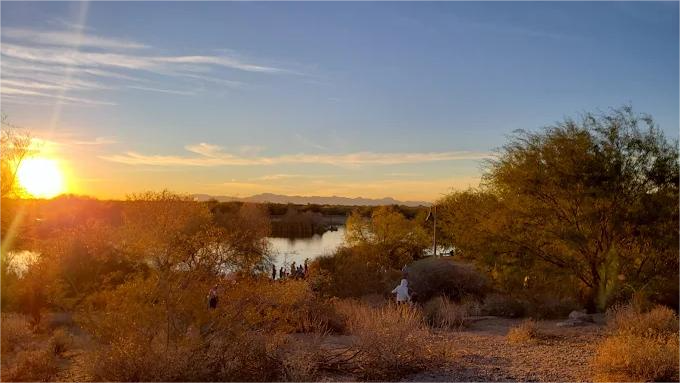 Sunset over a lake surrounded by desert vegetation, with a person standing in the distance.
