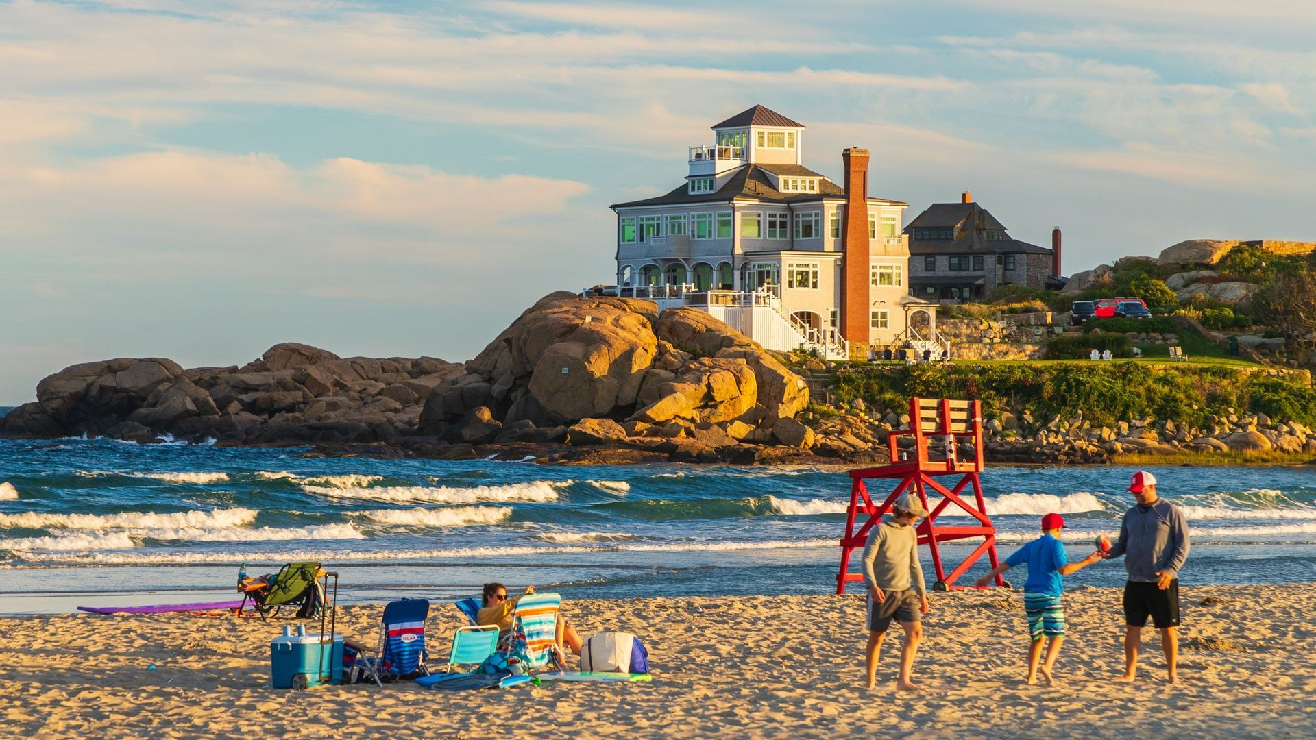Beach scene with ocean waves, a rocky coastline with buildings, and people on the sand.
