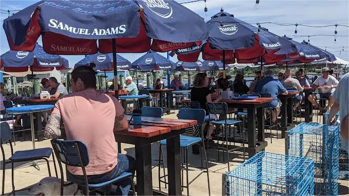 Outdoor dining area with blue Samuel Adams umbrellas, tables, and people enjoying food/drinks.