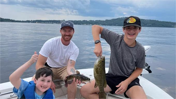 Three people on a boat fishing. A boy holds up a fish, two others show thumbs up. Lake background.