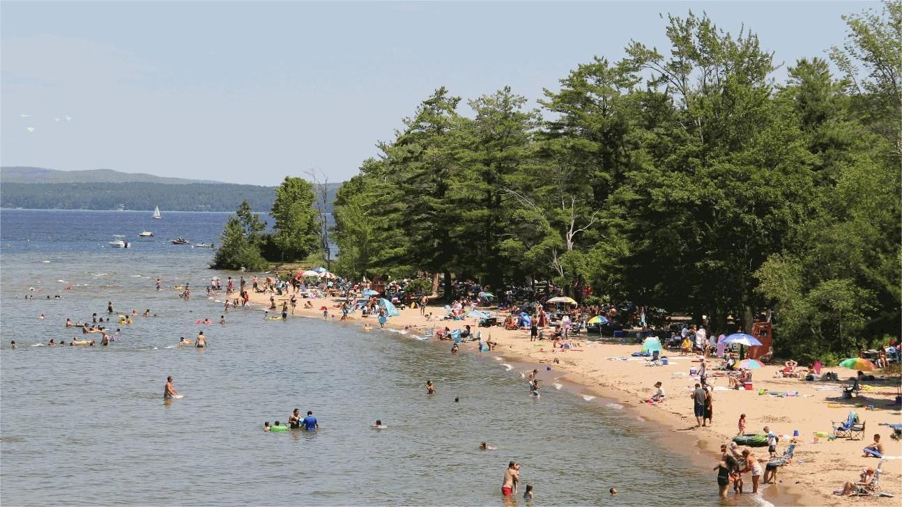 Beach scene with many people swimming and sunbathing; trees line the shore.