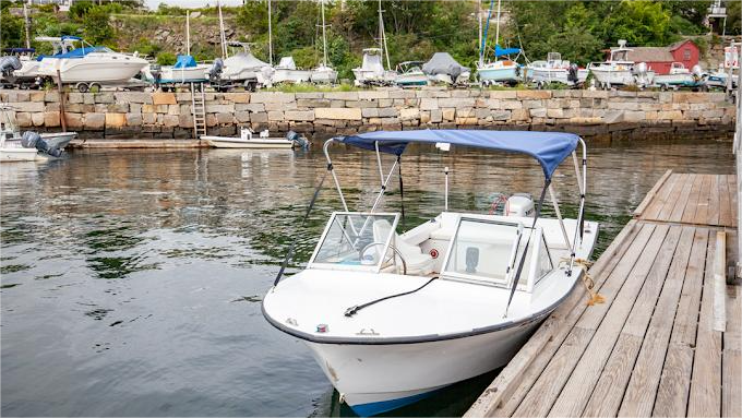 White motorboat with blue canopy docked at a wooden pier. Harbor with other boats in background.