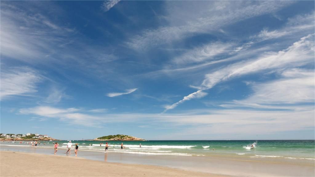 Beach scene with blue sky, white clouds, people near water.