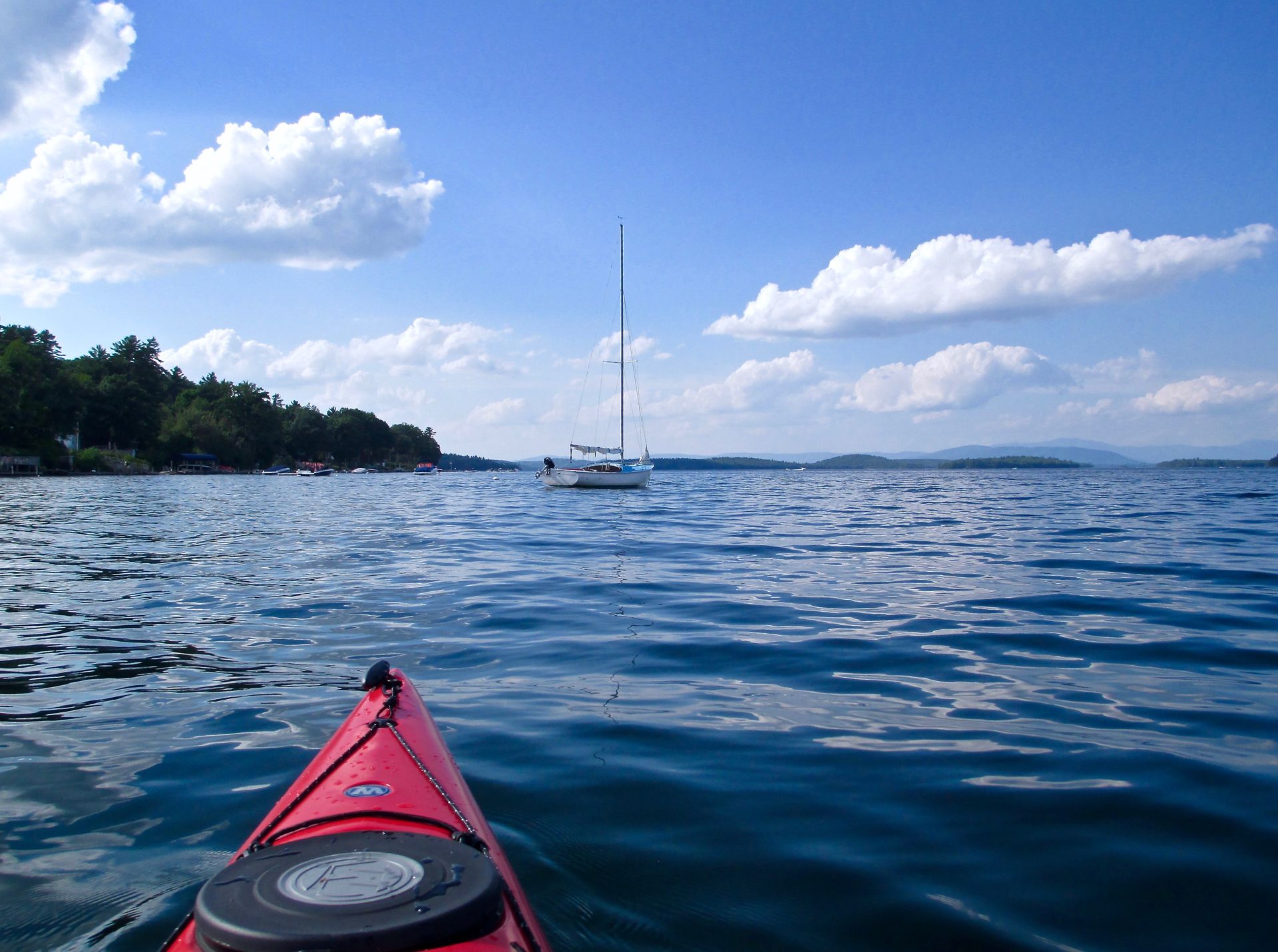 Red kayak on blue water, sailboat in the distance, clear sky with clouds.