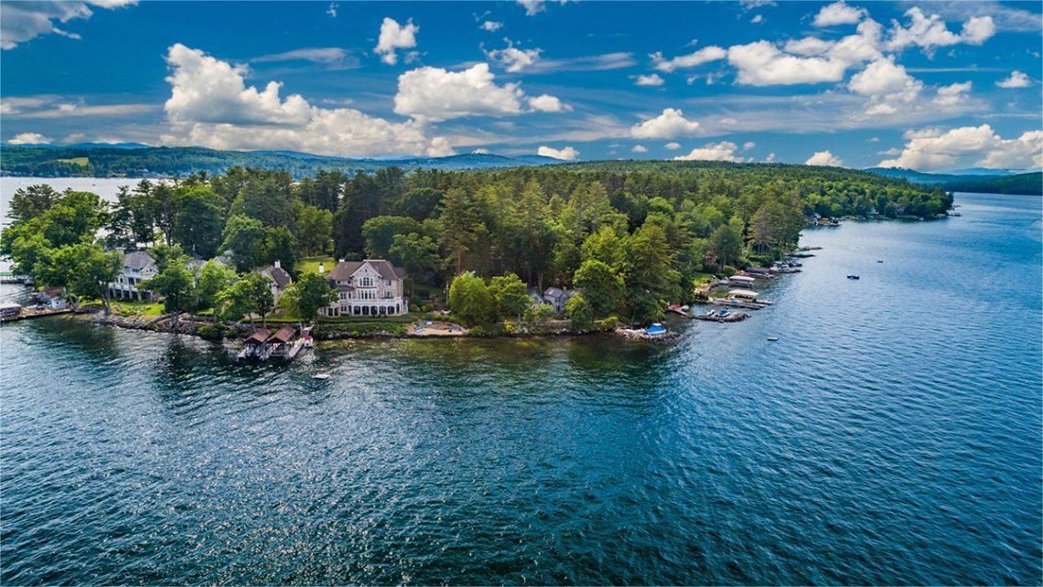 Aerial view of a wooded island surrounded by blue water, with several houses and docks visible.