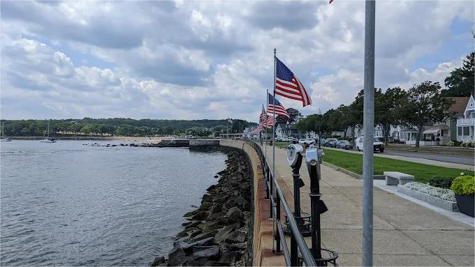 Waterfront with American flags waving; rocky breakwater, sidewalk, buildings under cloudy sky.