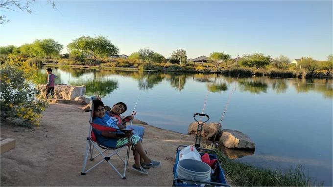 Two people fishing at a lake; one seated in a chair, holding a fishing rod, near a wagon with supplies.