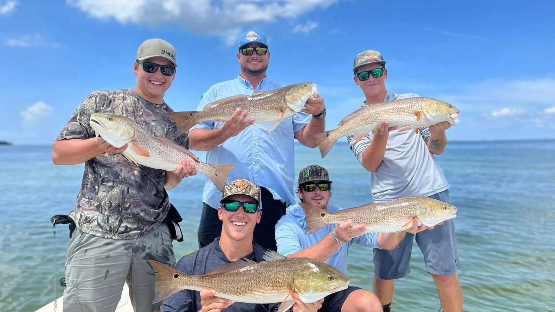 Six people on a boat holding up large redfish on a sunny day with blue water and sky.