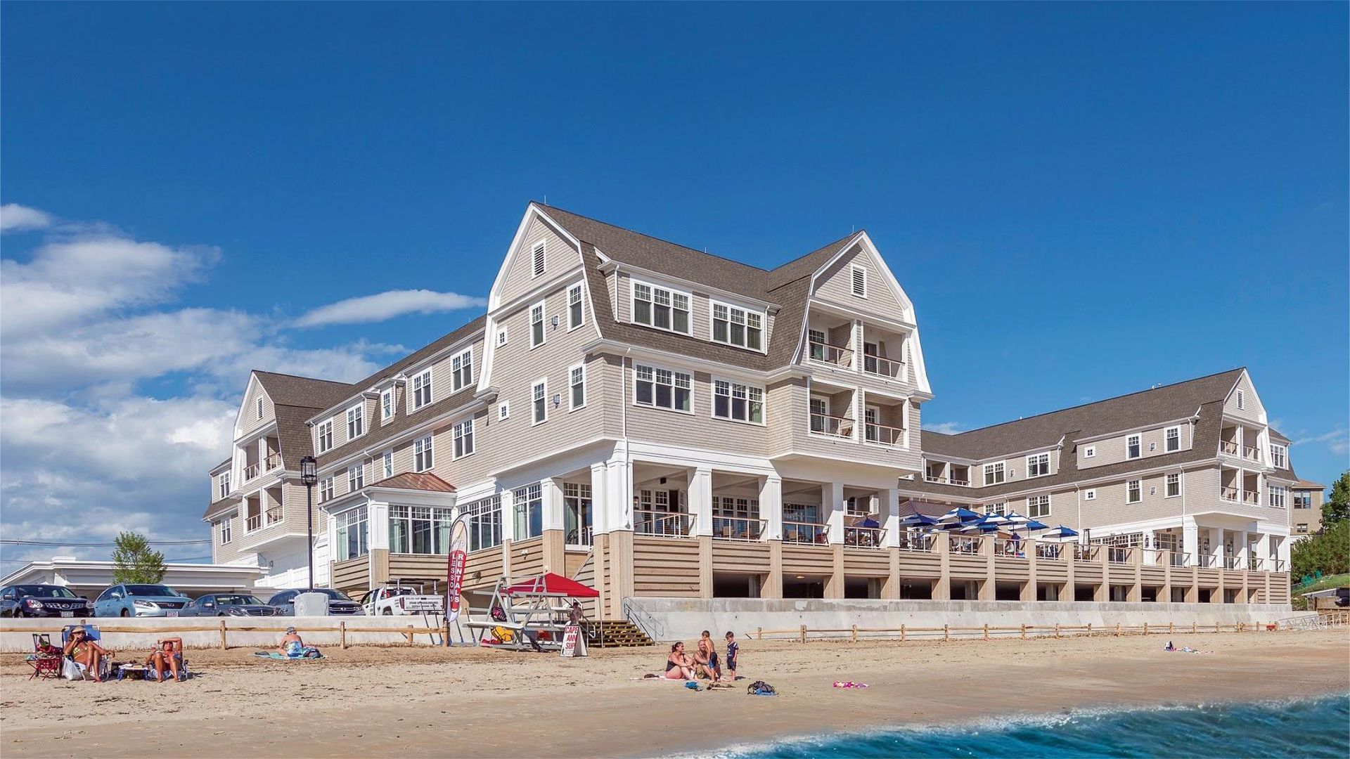Hotel on a beach with light-colored siding, a wooden deck, and people lounging on the sand under a blue sky.