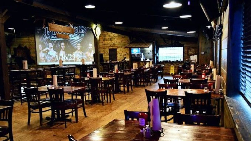 Interior of a rustic bar with wooden tables and chairs. Two large screens display images. Dim lighting.