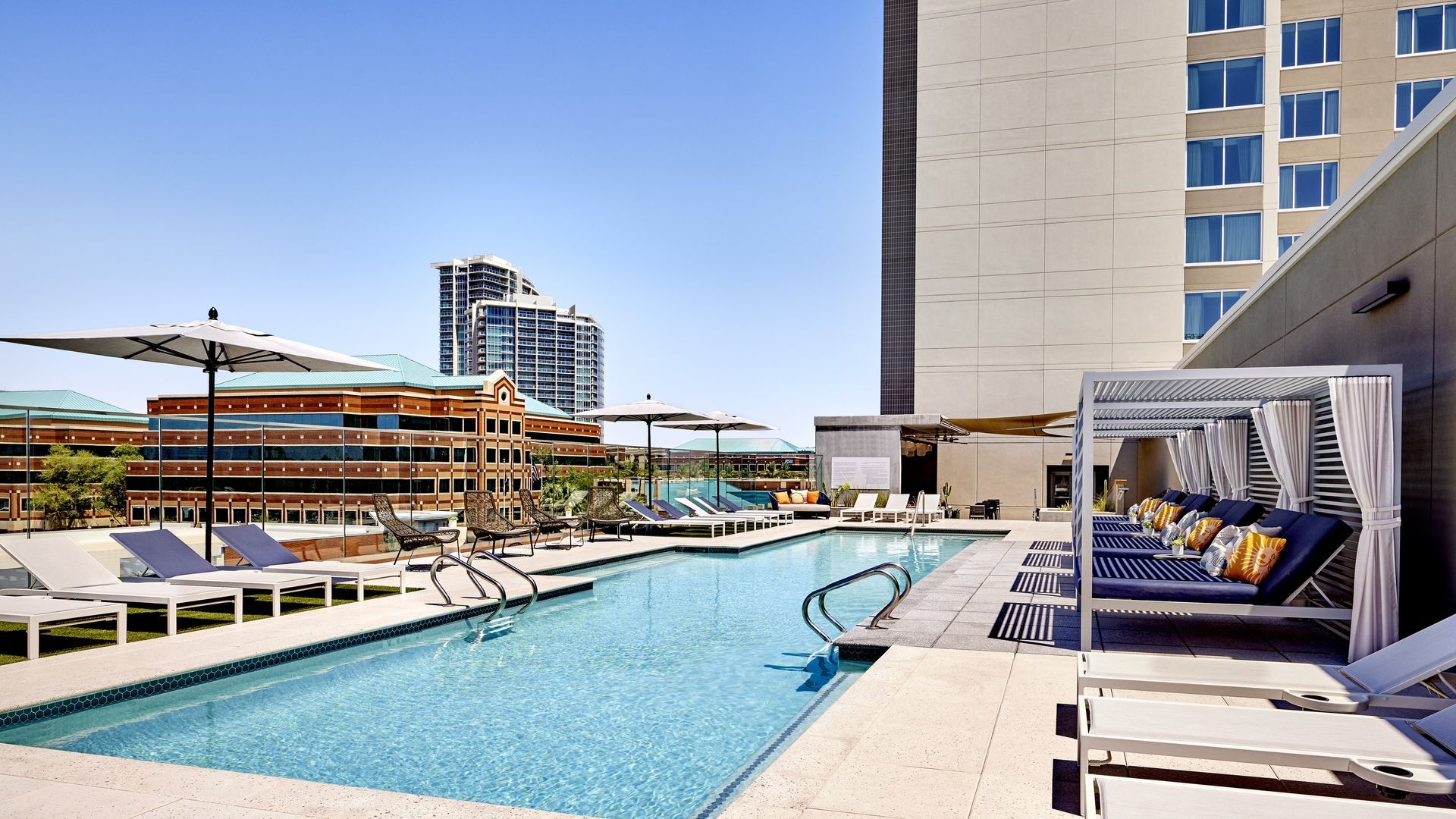 Rooftop pool with chaise lounges, cabana, and umbrellas. Buildings and blue sky in background.