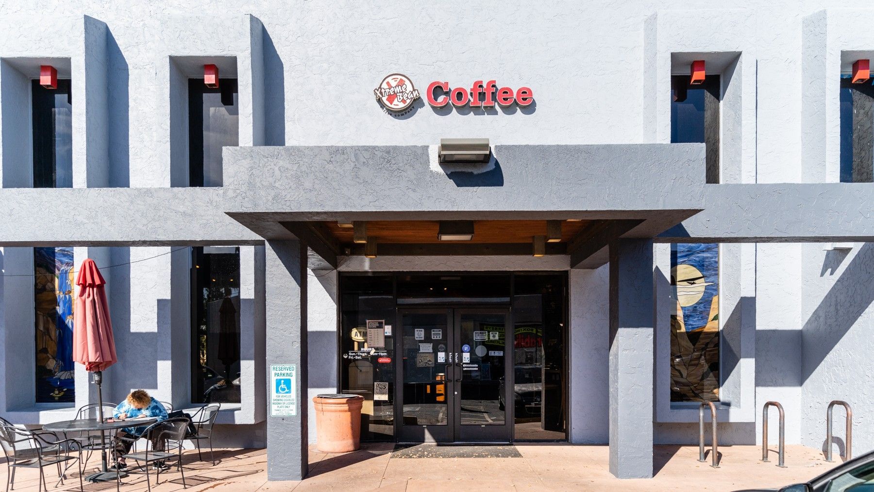 Exterior of coffee shop with red sign, gray facade, and a person seated outside.