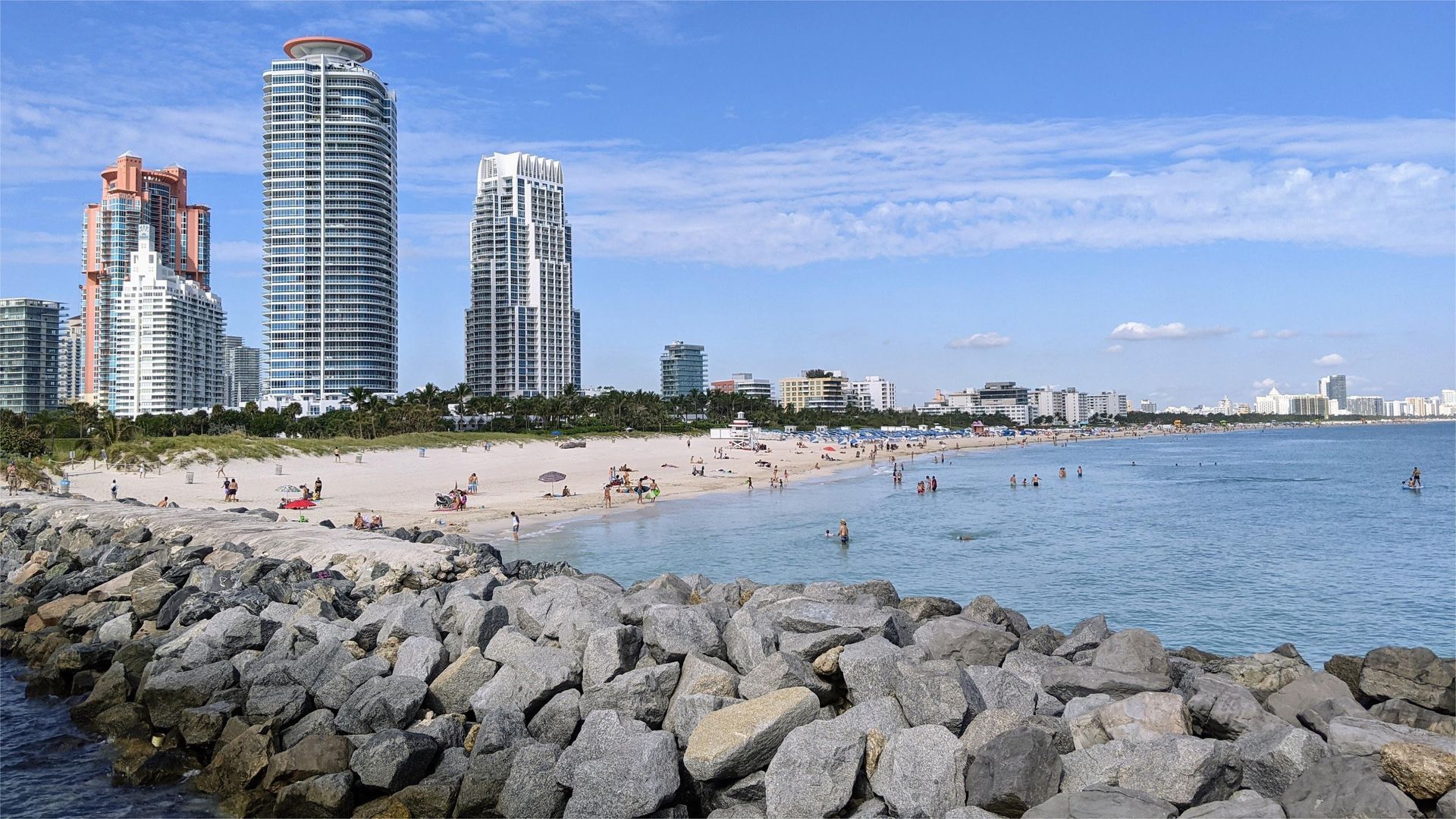 Beach scene with ocean, sand, and buildings. People on the beach, blue sky.