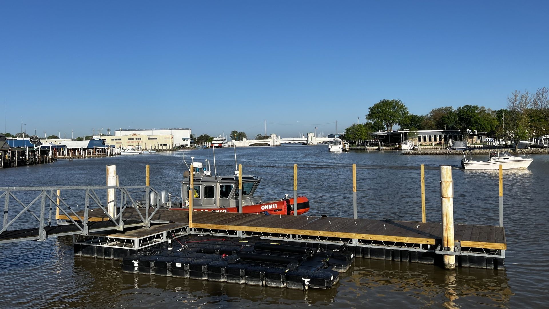 A dock on water with a boat, buildings, and trees under a clear blue sky.