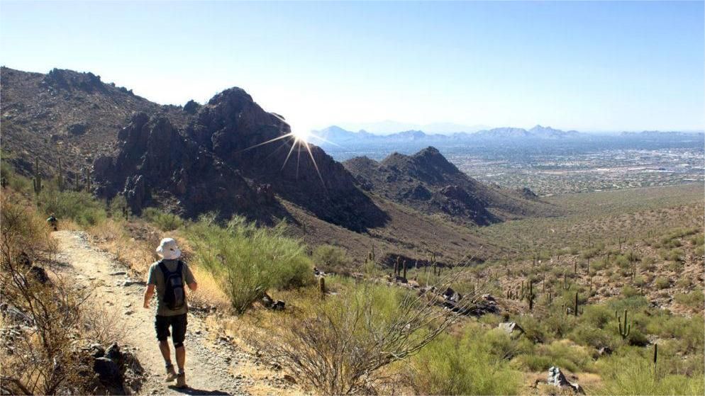 Person hikes on a desert trail, overlooking mountains and a distant city under a sunny sky.