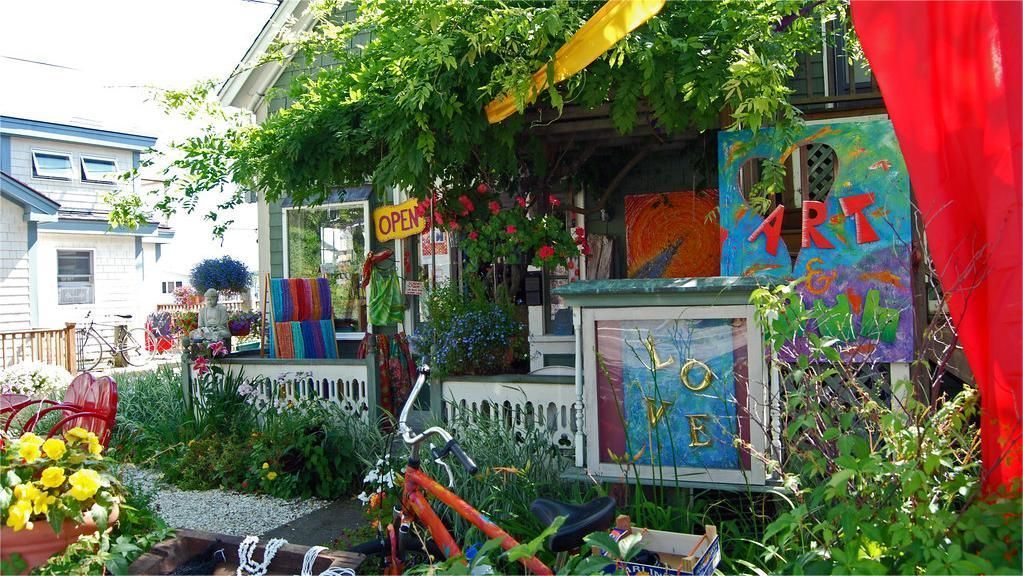 Quaint art shop exterior, lush greenery, colorful artwork displayed, open sign visible.