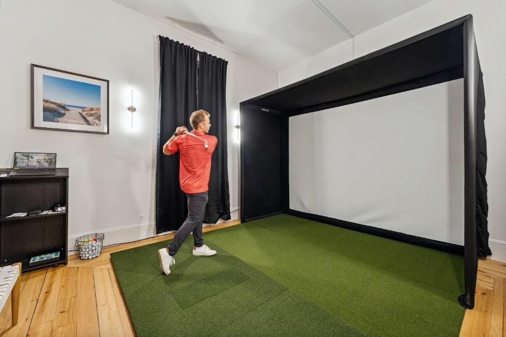 Person in a red shirt stands in a room with a golf simulator screen and green turf.