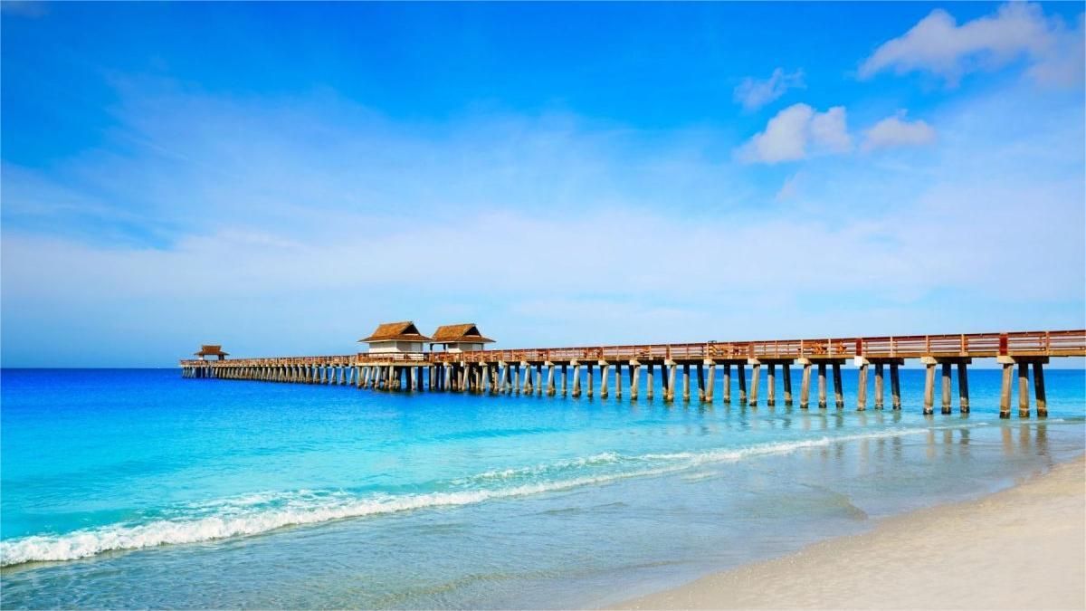Wooden pier extends into blue ocean under a bright sky. Waves wash onto the sandy shore.