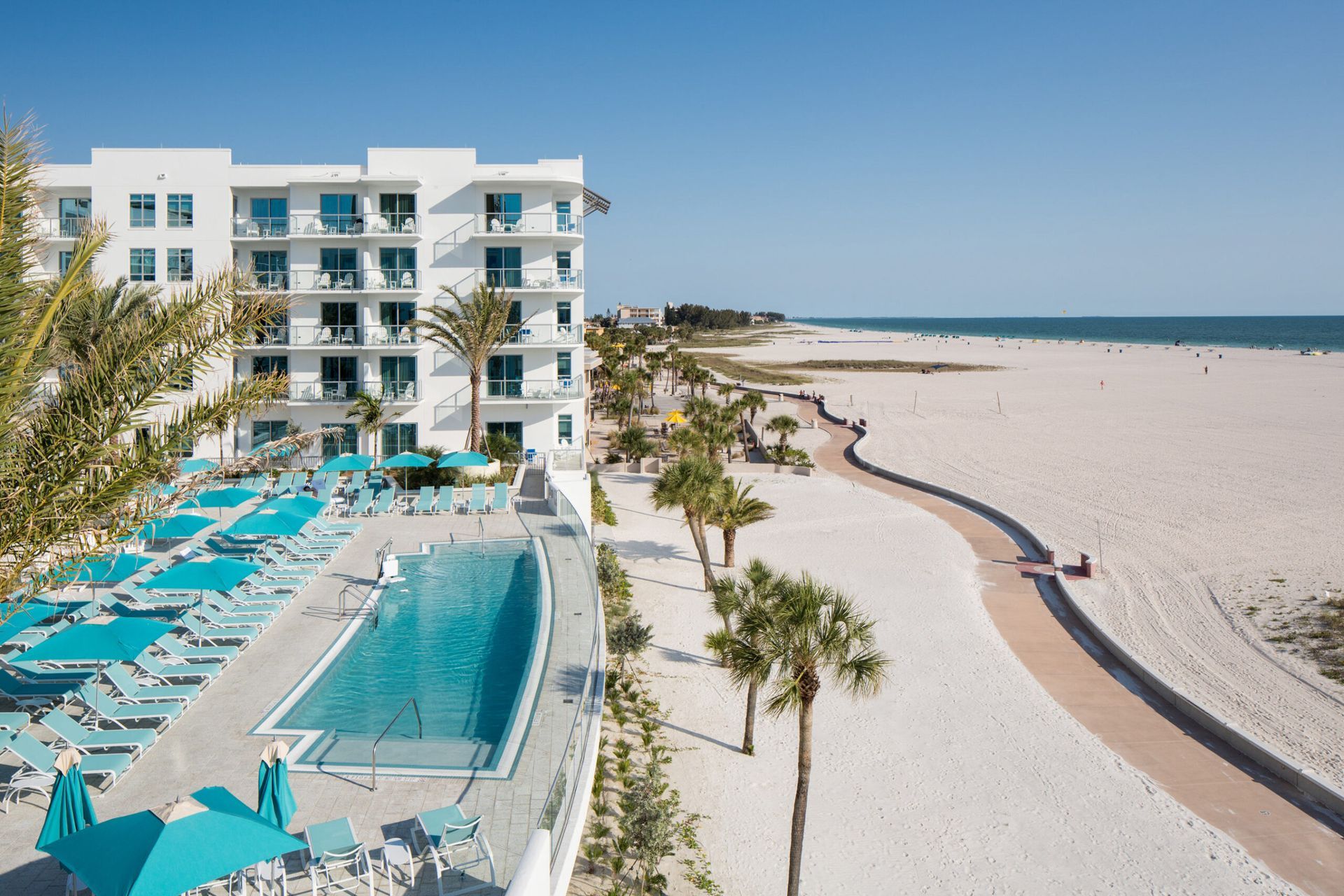 Hotel with pool and beach access. White building, turquoise accents, palm trees, blue sky and ocean.