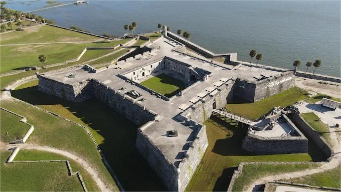 Aerial view of a star-shaped fort with surrounding earthworks, near water. Gray stone walls, green grass.
