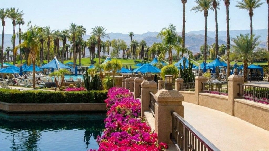 A sunlit resort pool area with blue umbrellas, palm trees, and a bridge lined with bright pink flowers.