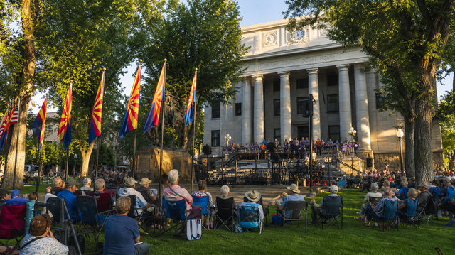 Crowd seated on lawn before a neoclassical building with rainbow flags and a stage during an outdoor event