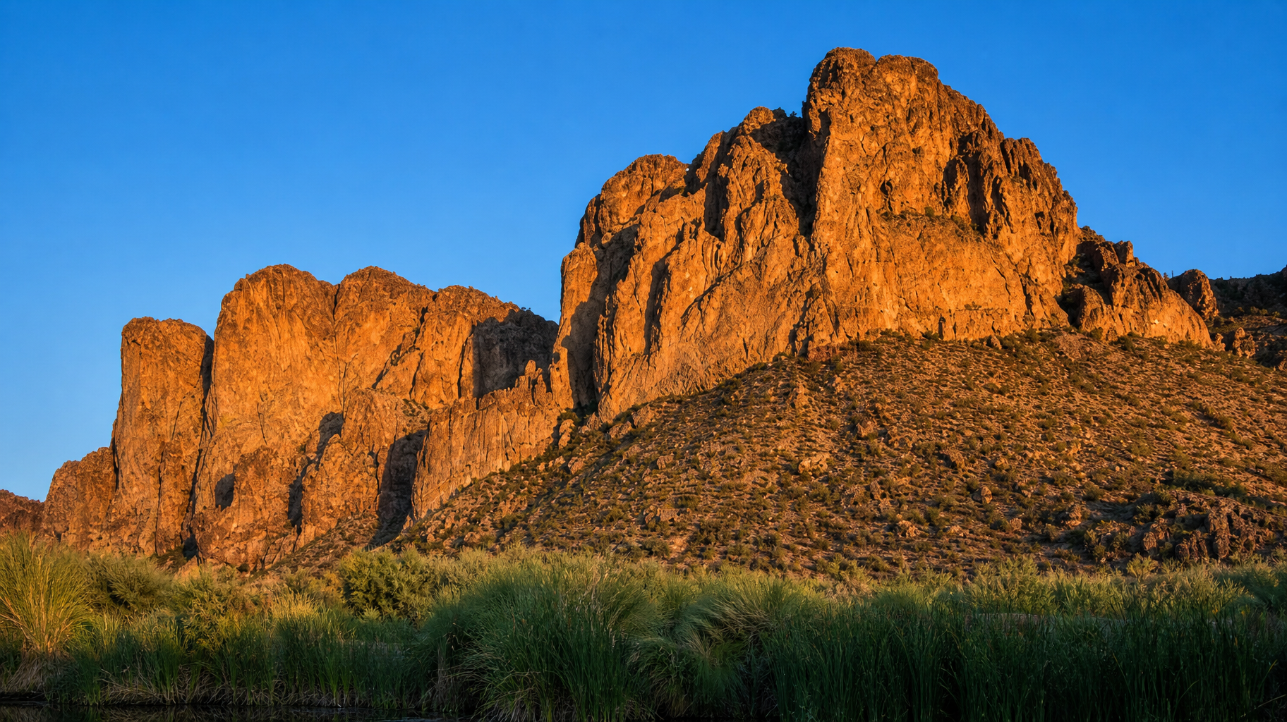 Sunlit orange rock formations above green grass under a clear blue sky