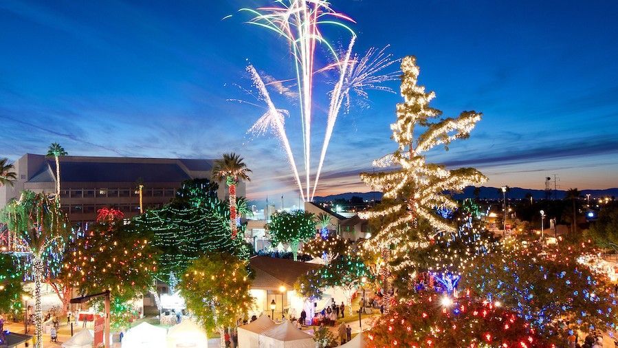 Fireworks explode over a town square with a lit Christmas tree and buildings at night.