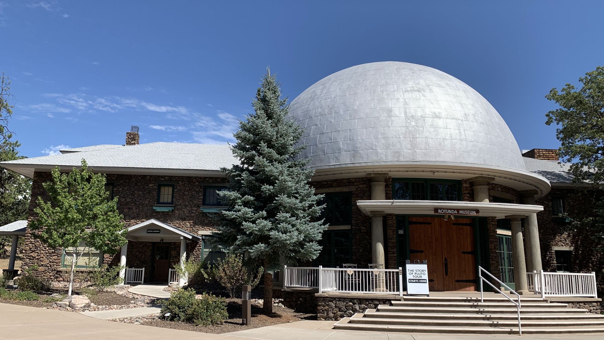 Building with a large silver dome and a wooden entrance, trees in front.
