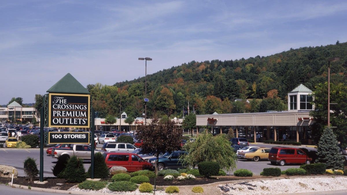 The Crossings Premium Outlets sign sits in the foreground of a shopping center with a parking lot and tree-covered hill.
