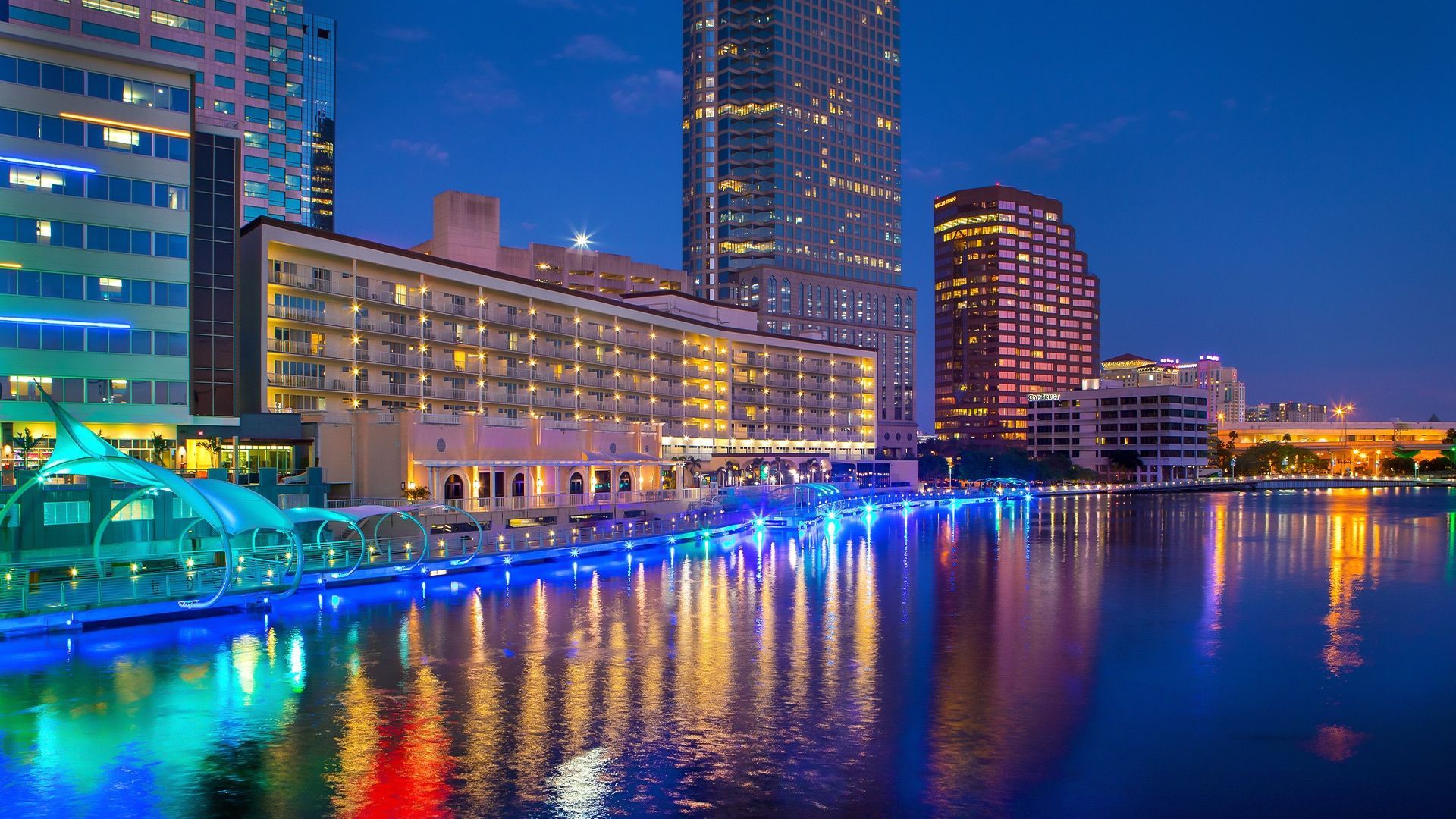 City skyline at dusk reflecting in a river, illuminated buildings and water feature with blue and green lights.