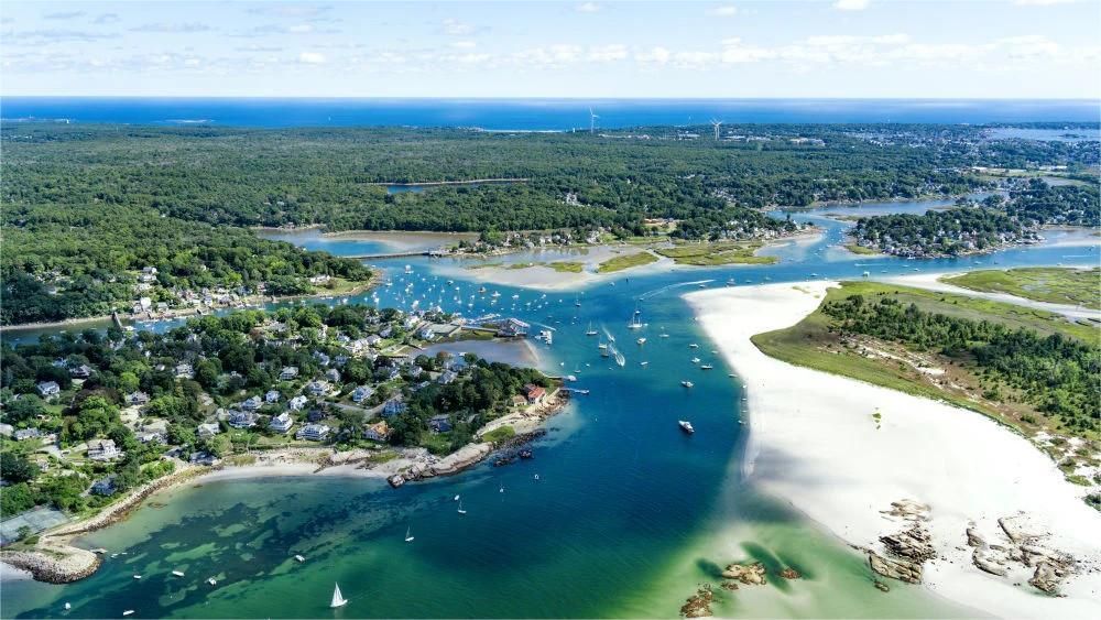 Aerial view of a coastal inlet with boats, sandy beaches, green trees, and blue water under a clear sky.