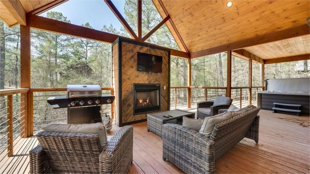 Screened-in porch with fireplace, grill, seating, and hot tub. Wood ceiling, forest view.