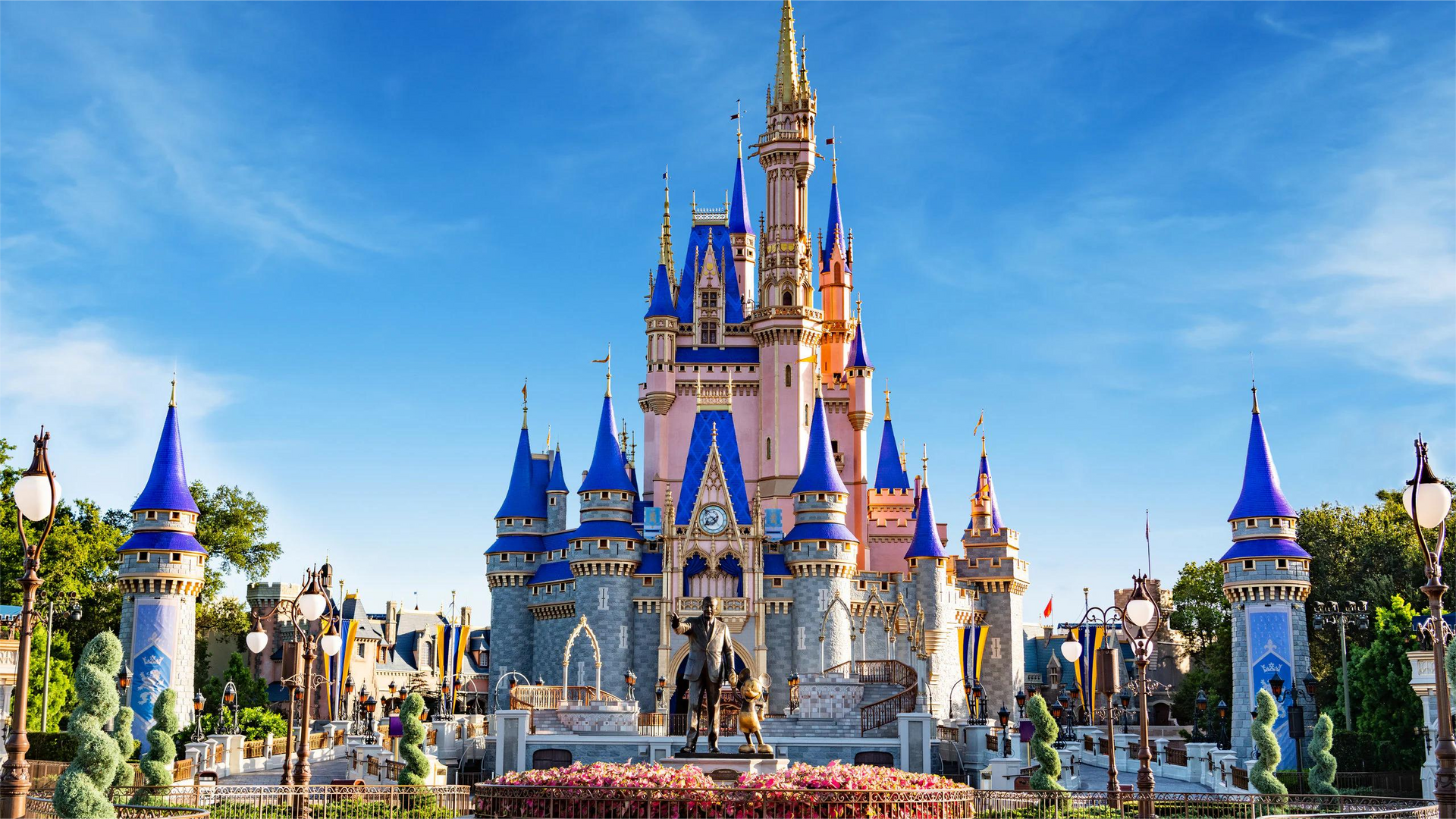 Cinderella Castle at Walt Disney World under a bright blue sky with a bronze statue of Walt Disney and Mickey Mouse.