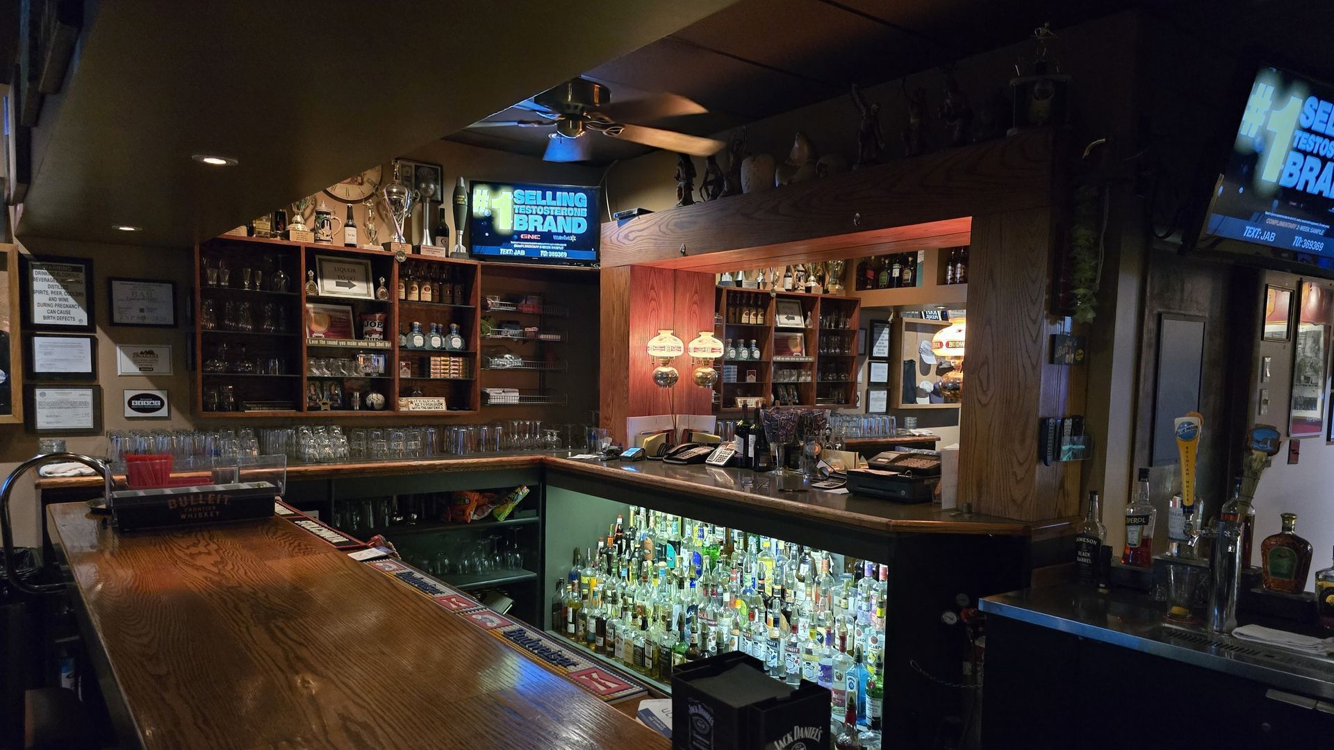 Interior view of a dimly lit bar, showcasing a wooden bar, shelves of liquor, and TVs.