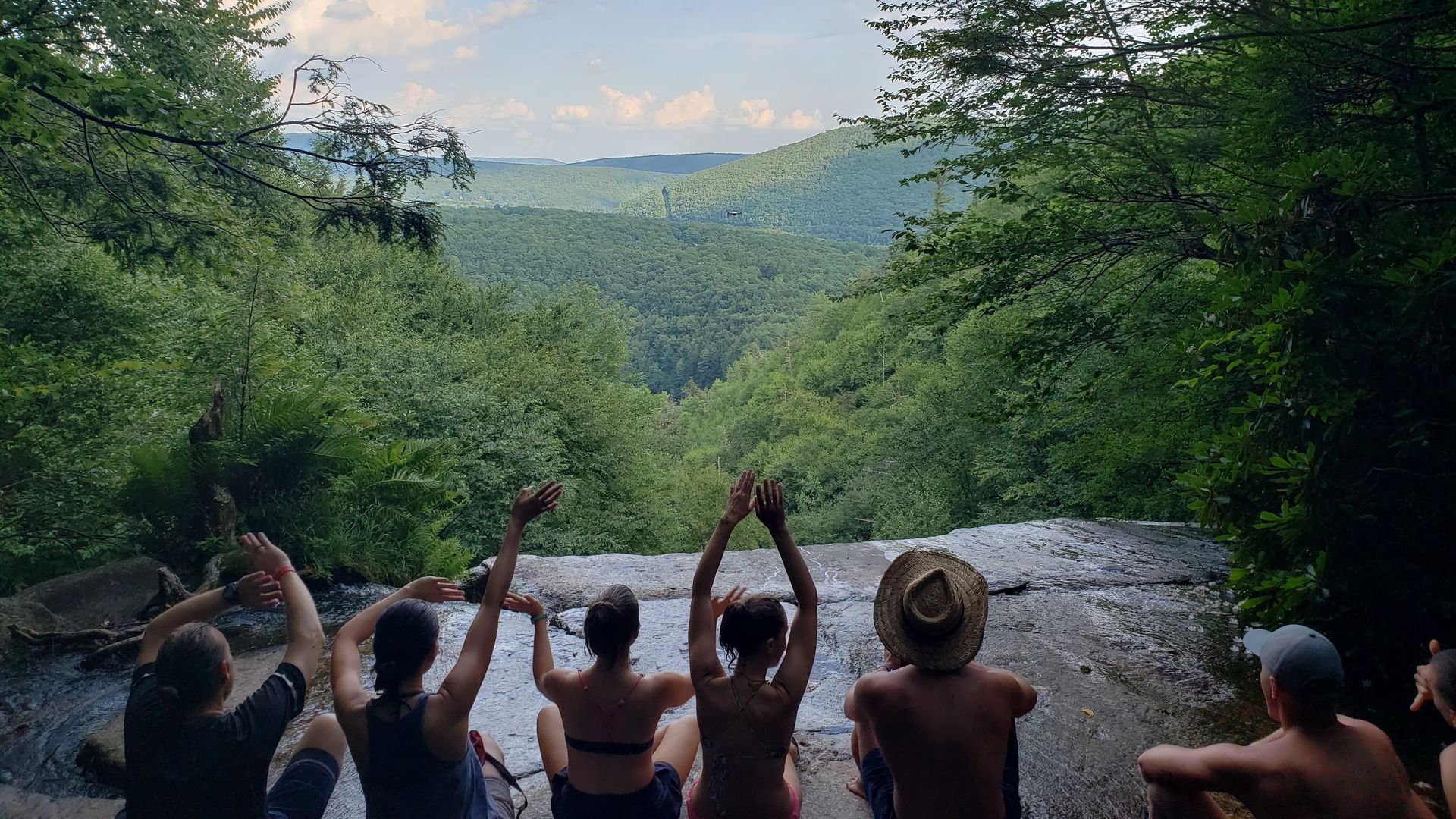 People sitting on a waterfall edge, arms raised, overlooking a green valley. Sunny day.
