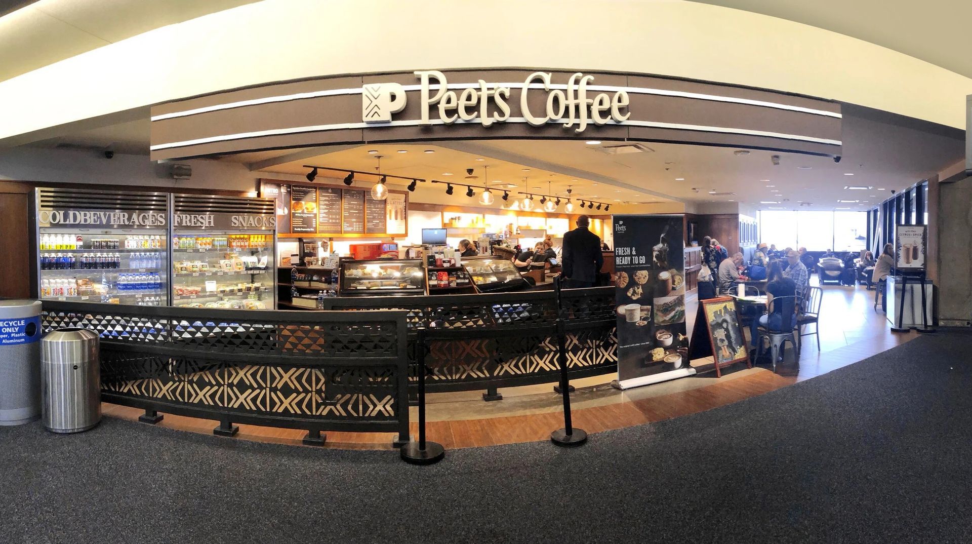 Peet's Coffee shop in an airport. People stand at counter and sit at tables. Gray floor and white sign.
