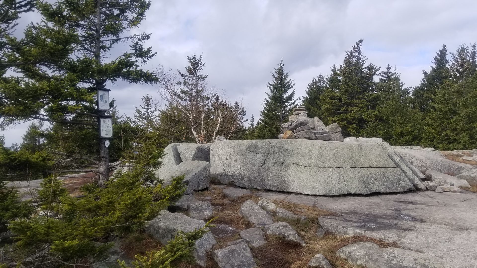 Rocky mountaintop with stone structure, cairn, and signs, surrounded by trees under a cloudy sky.